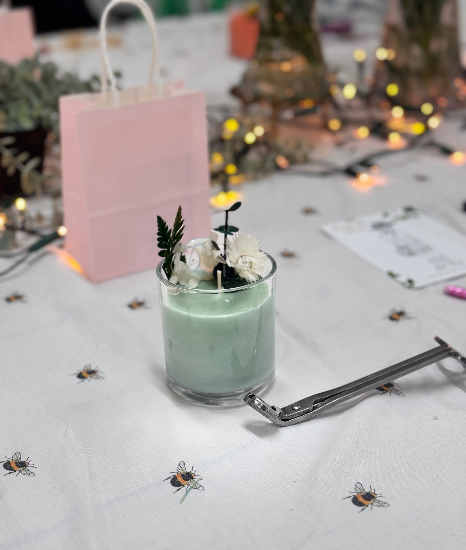 Green candle in a clear glass jar with green and cream dried flowers on top for decoration. A silver wick trimmer is on the side of the table and the background which is out of focus has fairy lights, a pink giftbag and flowers in vases.